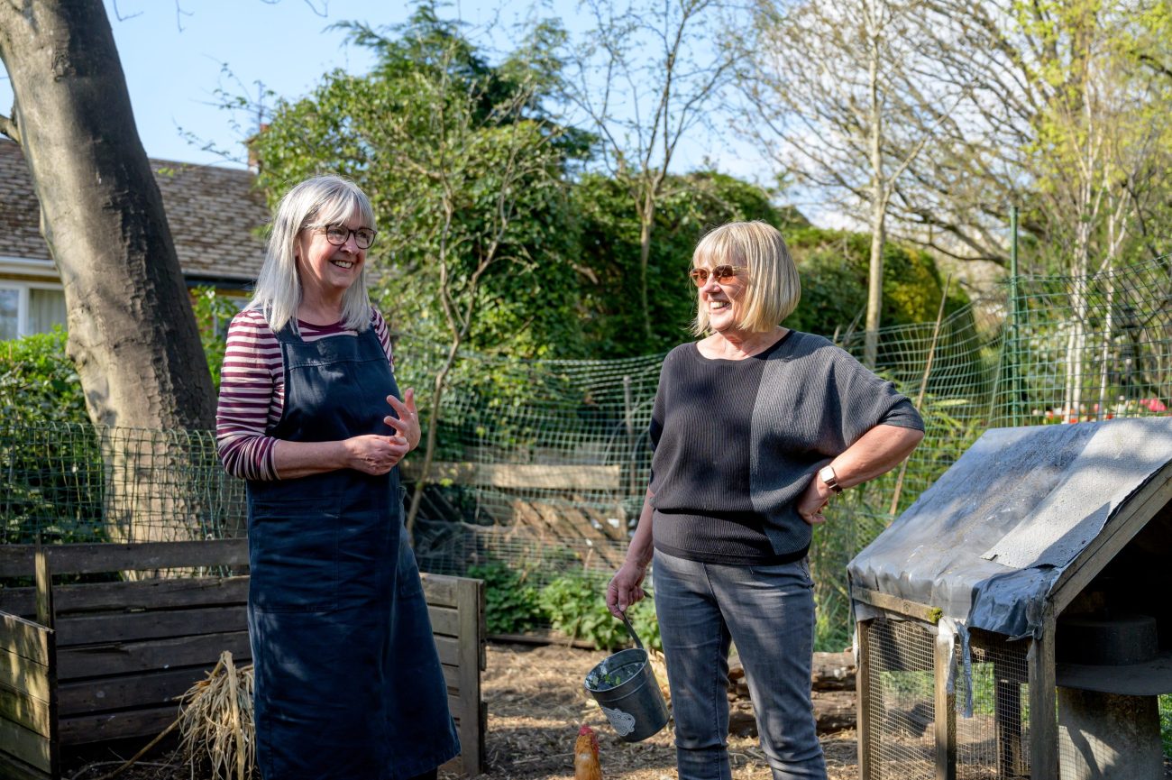 Two white women with grey hair stand in a sunny community.