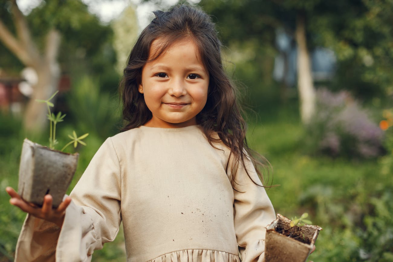 A young girl smiles while holding potted plants in a lush, sunny garden.